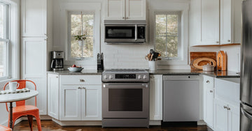 a bright kitchen with white cabinets, stainless appliances, large windows, and an orange chair by a round table.