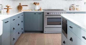 A kitchen with dark cabinets, a cream stove, white tile backsplash, wall shelves, plants, and wood flooring.